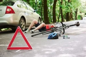 Injured cyclist lays on road next to car and overturned bicycle with a hazard triangle in the foreground.