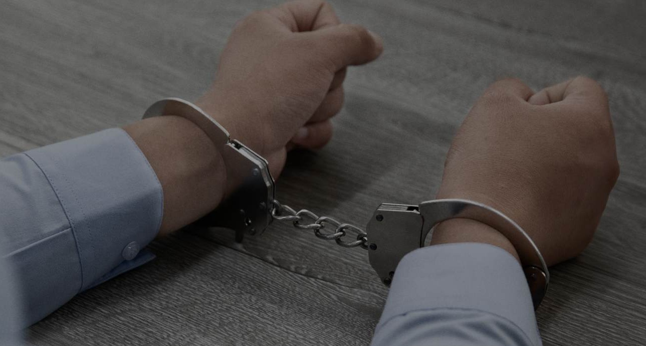Closeup of business man in button-down shirt wearing handcuffs with his arms resting on table.