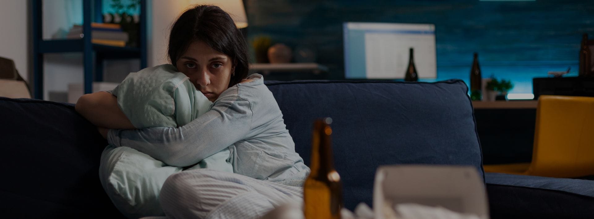 Fearful, young indigenous woman who was a sex crime victim hugs pillow in somber living room with empty alcohol bottles in background.