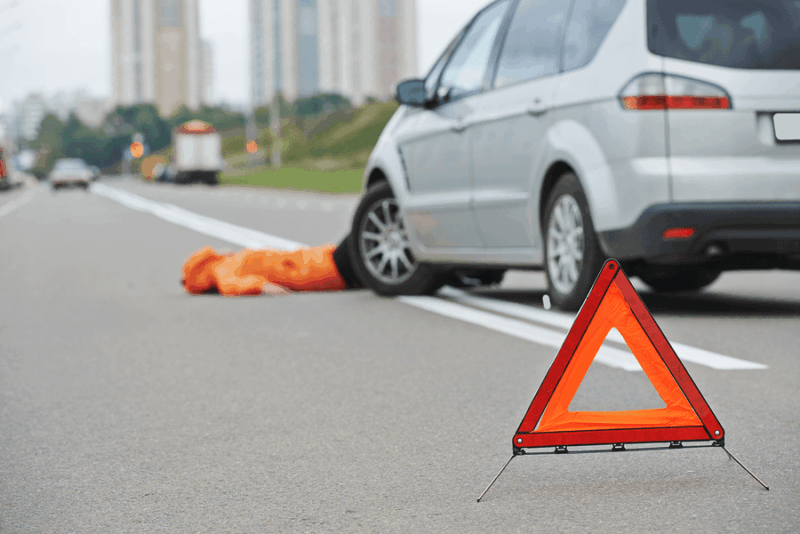 Image of minivan stopped in the road with a pedestrian laying on the pavement and a hazard triangle in the foreground.
