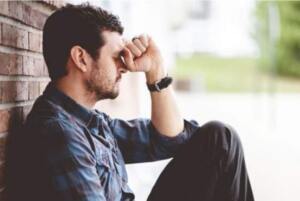Vulnerable man in plaid shirt holds his fist against his head as he sits next to a brick building.