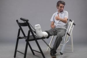 Injured man gestures a thumbs up with his broken leg propped up on a chair and his crutch resting beside him.