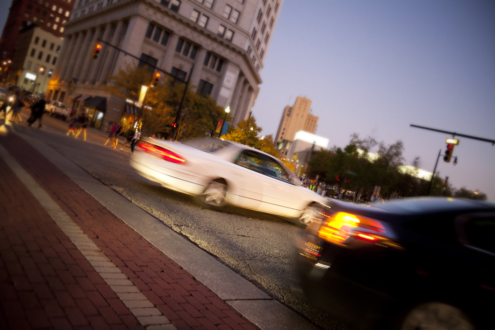 Two cars speeding through the streets of Grand Rapids, Michigan. Pedestrians and multiple buildings can be seen in the background.