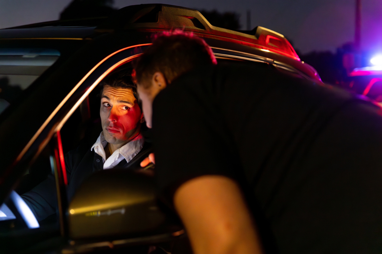 A police officer is talking to a young adult male driver pulled over at night. The driver has a guilty expression.