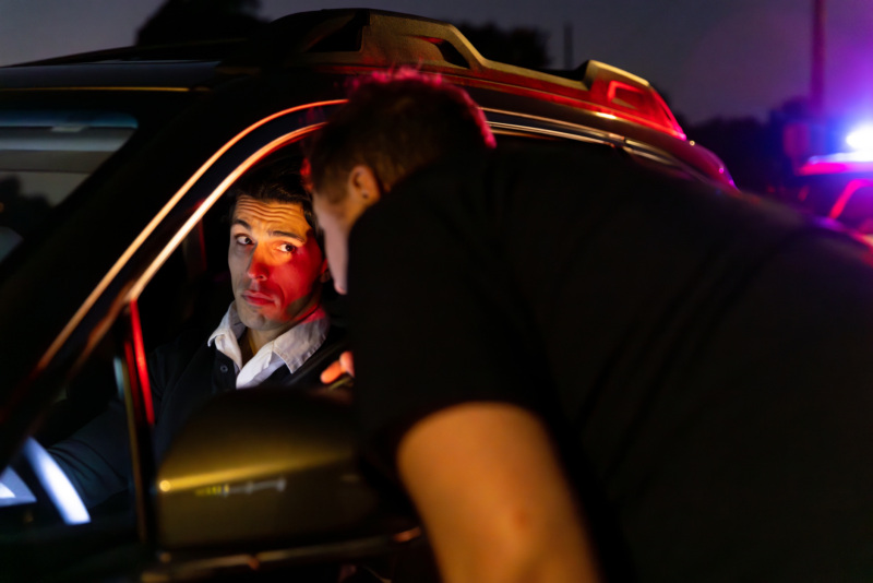 A police officer is talking to a young adult male driver pulled over at night. The driver has a guilty expression.