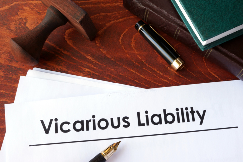 A stack of documents, the top one labelled, “Vicarious Liability”. A fountain pen is uncapped atop the documents, with a stamp and 2 books nearby on the desk.